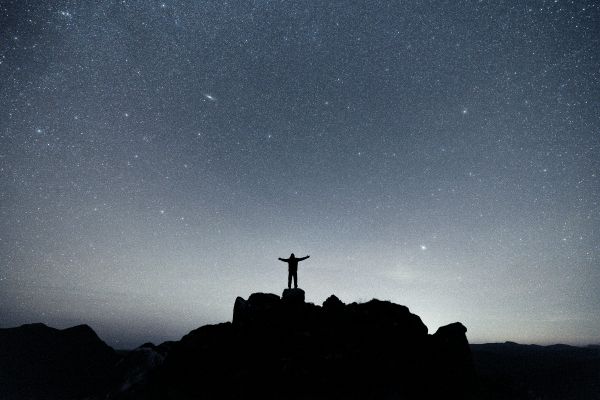 a person standing on a mountain top with arms raised under a starry night sky, wondering is evolution true or did God create it all